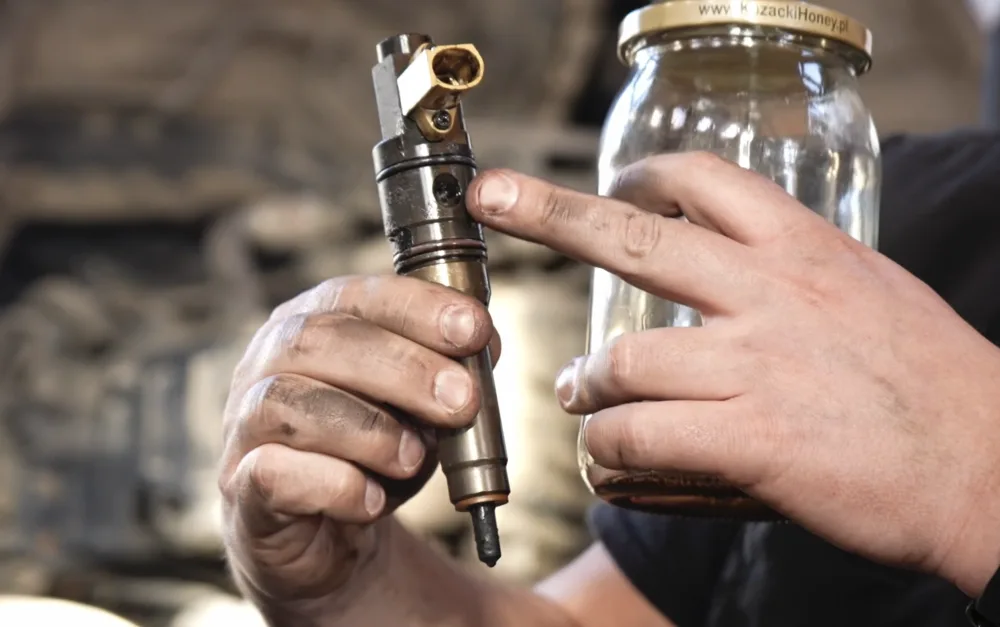 Diesel injector with carbon deposits on the nozzle tip held in a mechanic's hands next to a jar of fuel