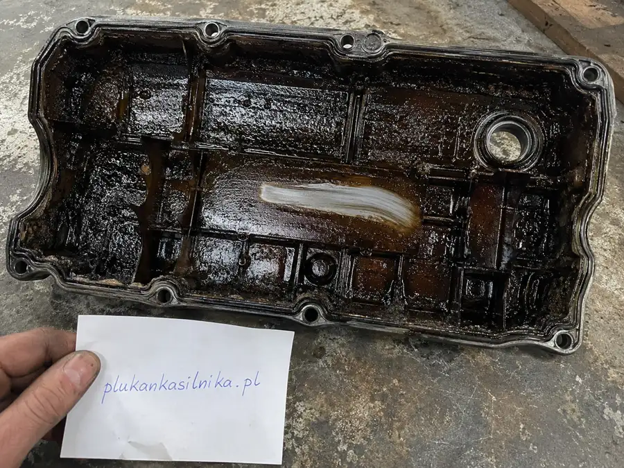 Underside of a removed rocker cover coated with dark deposits, with a wiped streak of clean aluminium