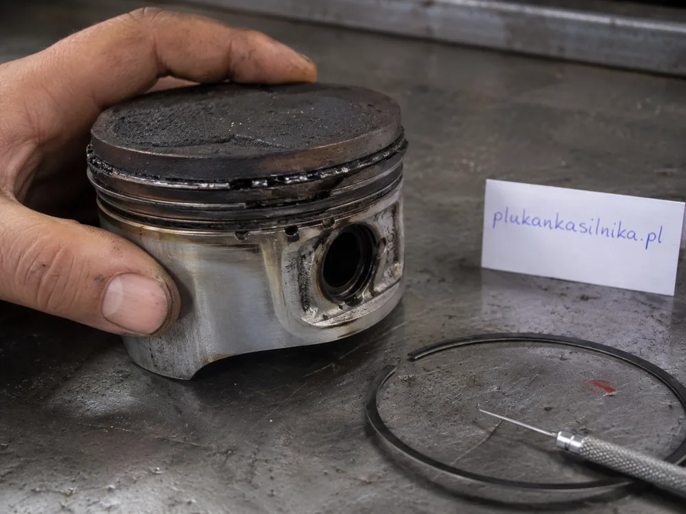 Piston removed from an engine, hard carbon visible in the ring grooves, with a piston ring and pick tool beside it